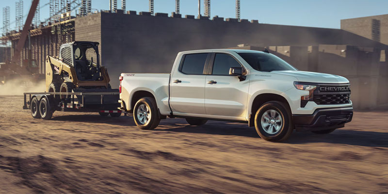 White Chevrolet Silverado towing a flatbed trailer carrying construction equipment across a dusty job site in Haskell, Texas, with unfinished buildings and steel frameworks in the background.