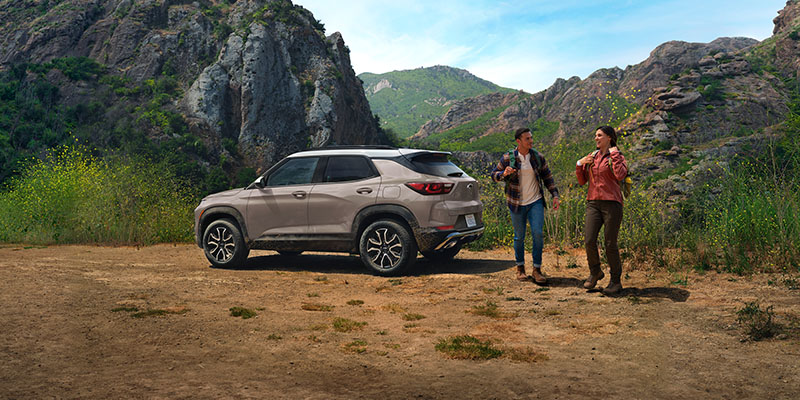 A beige compact SUV parked in a mountain valley with two hikers standing nearby on a scenic trail.